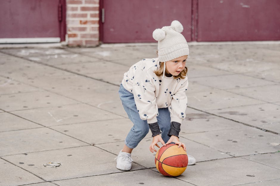 A young child plays with a basketball on a concrete court, dressed warmly for a chilly day.