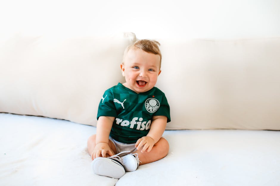 Cute baby smiling while sitting on a sofa wearing a green sports jersey indoors.