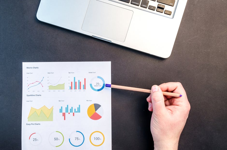 Hand holding pencil reviewing colorful data charts on desk with laptop.