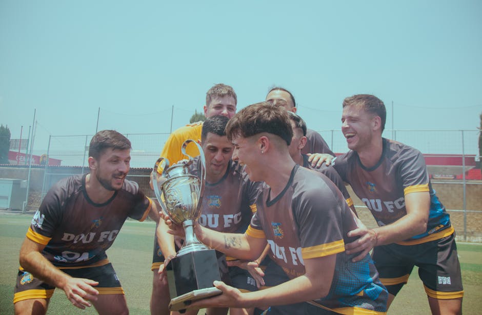Soccer team joyfully celebrating their victory on a sunny pitch holding a trophy.