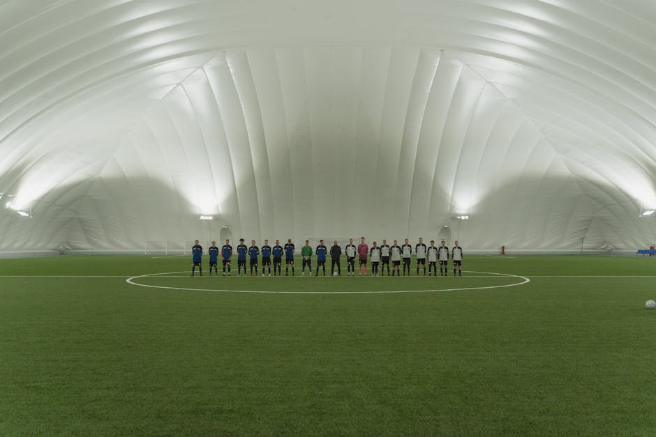 Two soccer teams lined up on an indoor artificial turf field under dome lighting.