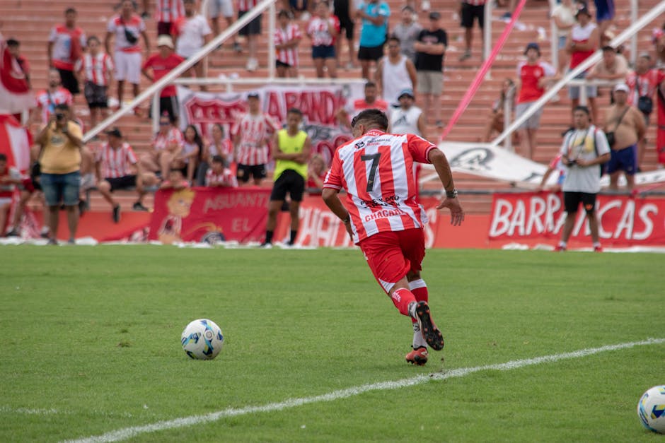 A soccer player in red and white stripes plays during a match, with fans watching from the stands.
