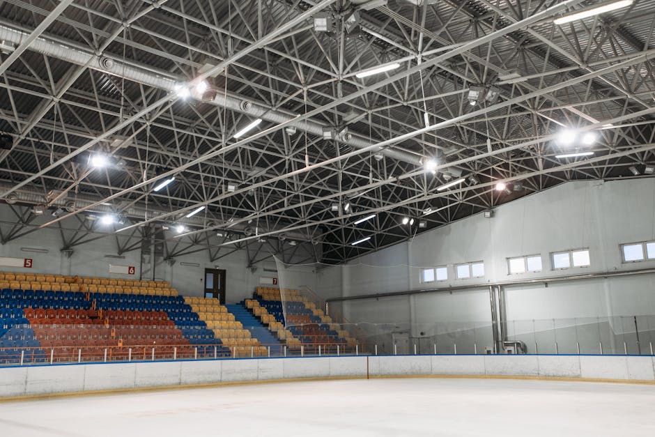 An empty indoor sports stadium with ice rink, colorful seating, and steel ceiling structure.