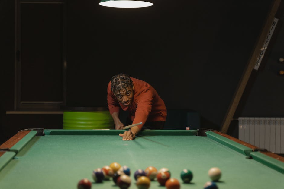 A person skillfully aiming a pool cue on a billiards table under dim lighting.
