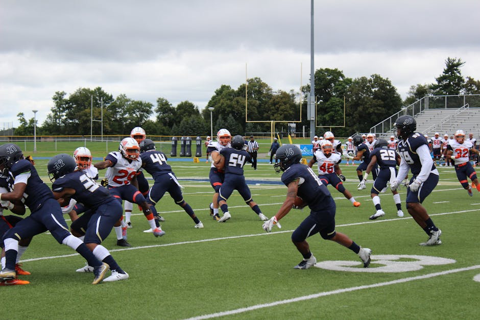 Football players in action during a competitive match on a green field under cloudy skies.