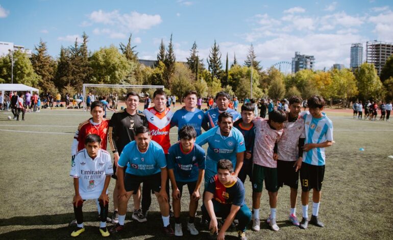 A youth soccer team poses on the field during a sunny day, with trees and city buildings in the background.