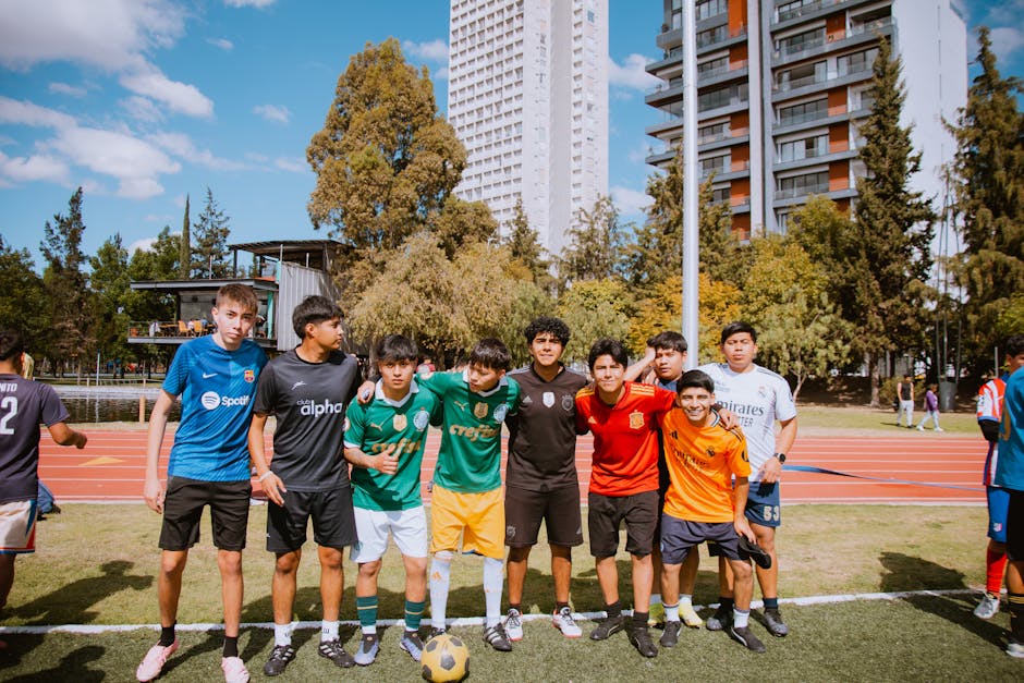 Teenagers enjoy a soccer game outdoors on a sunny day, featuring diverse jerseys and urban background.