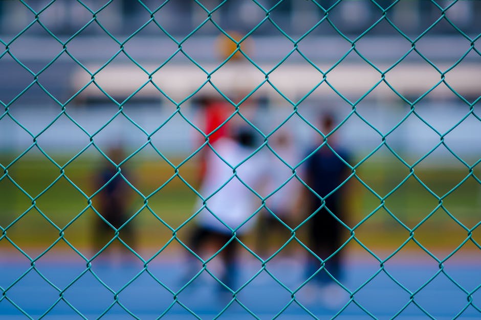 Blurred view of people playing sports behind a blue chain-link fence outdoors.
