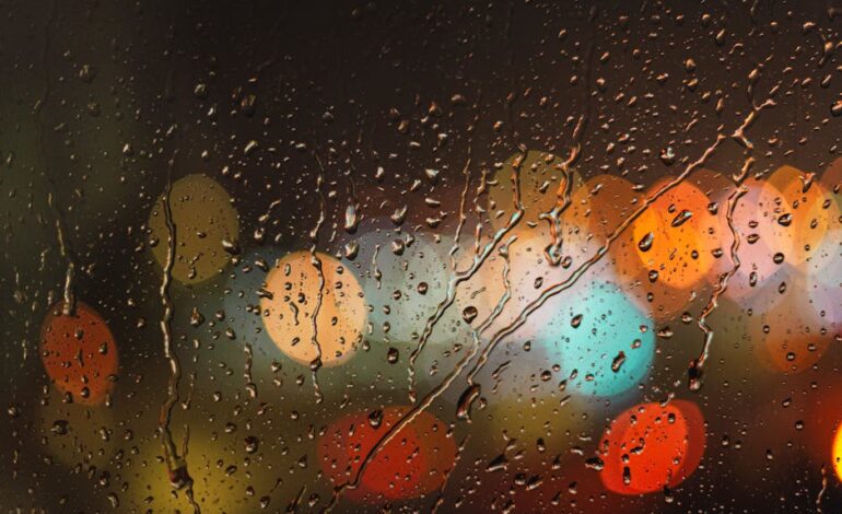Close-up of raindrops on glass with blurred colorful city lights in the background.