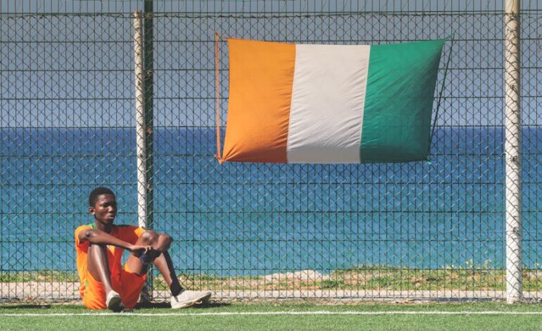Young athlete in orange jersey sitting by an Ivorian flag on a football field with sea view.