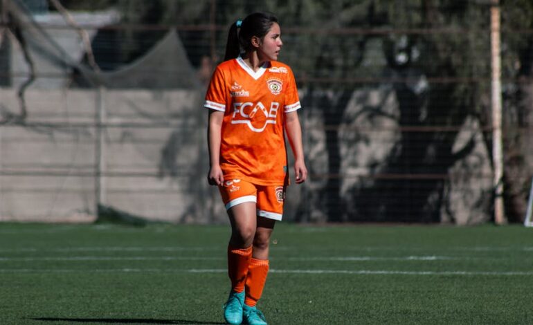 A female soccer player in orange uniform during a match on an outdoor field in Chile.