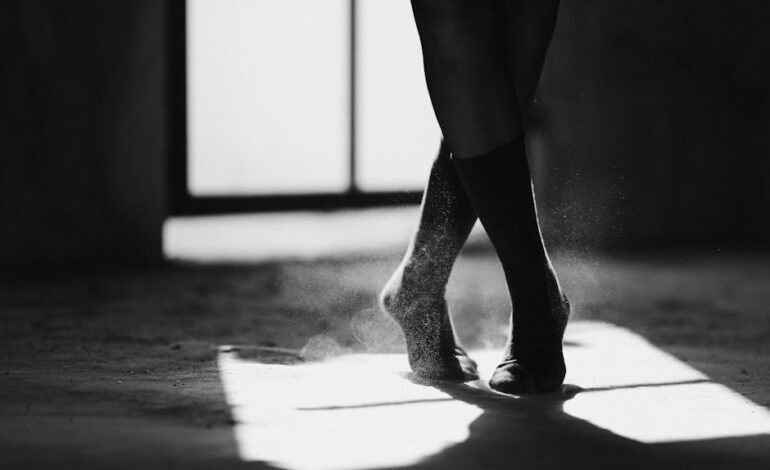 Black and white of crop anonymous female ballet dancer standing on dusty floor with crossed legs