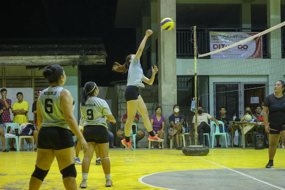 A competitive indoor volleyball game with women athletes jumping to spike the ball at night.