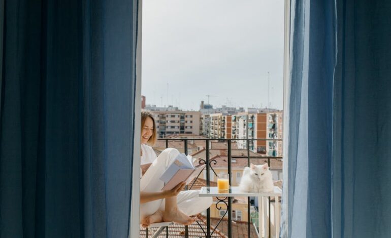A woman enjoys reading on her balcony with a white cat and a glass of juice.