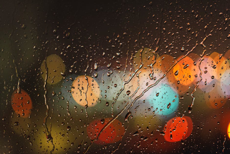 Close-up of raindrops on glass with blurred colorful city lights in the background.