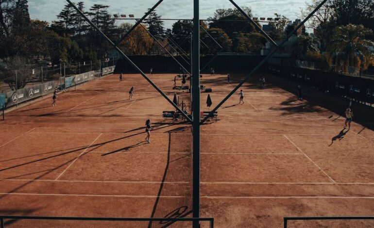 Tennis players compete on clay court with autumn foliage in the background.