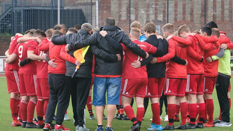 A soccer team huddles together on the field wearing red jerseys, showing unity and teamwork.