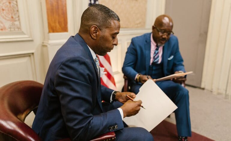 Two men in business attire discussing documents, American flag in background.
