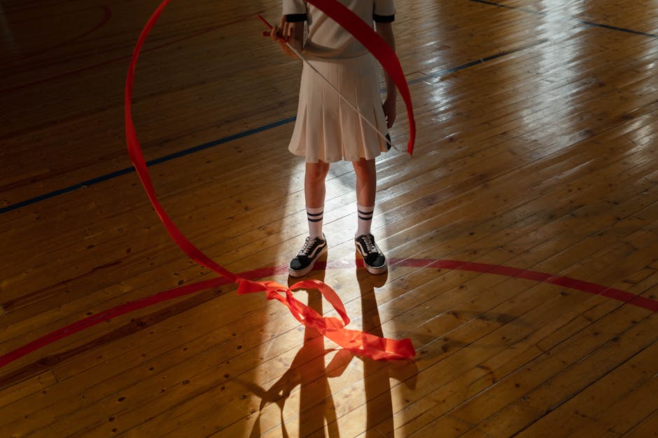 A young girl performing ribbon gymnastics indoors on a wooden floor.