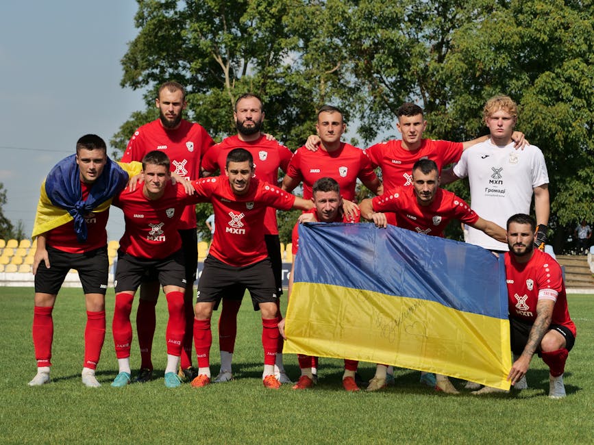 Soccer team in red jerseys posing with Ukrainian flag on a sunny day.