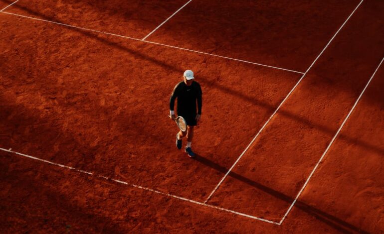 An overhead shot of a tennis player on a clay court during the day, with long shadows.