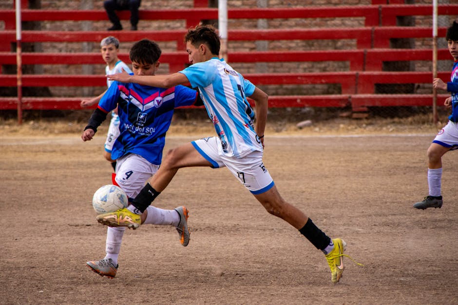Teen soccer players compete fiercely during a match on a dirt field with red bleachers.