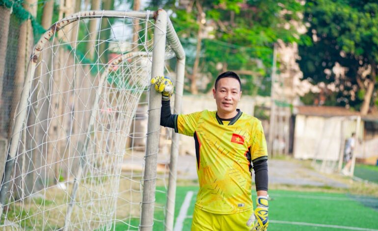Vietnamese soccer player in a yellow goalkeeper uniform posing by a goal on a sunny day.