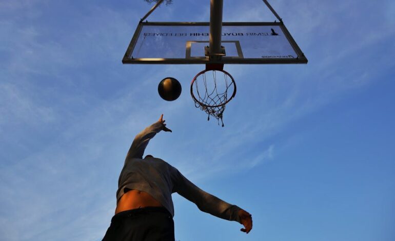 Athlete jumping to shoot a basketball with clear blue sky backdrop, captured from a low angle.