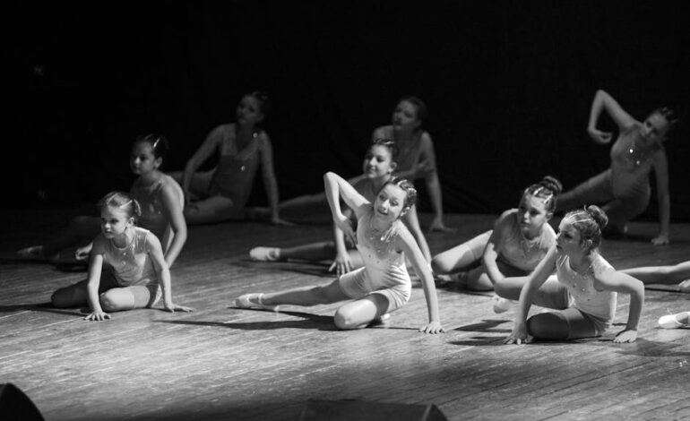 Young ballerinas performing a choreography on stage in a monochrome setting.
