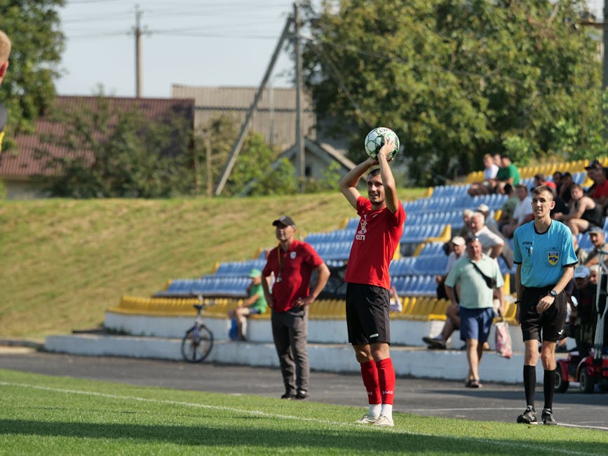 Soccer player prepares to throw in the ball during an outdoor match, with referee and spectators watching.