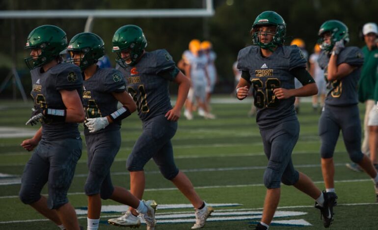 High school football team running on field during sunset, wearing green uniforms and helmets.