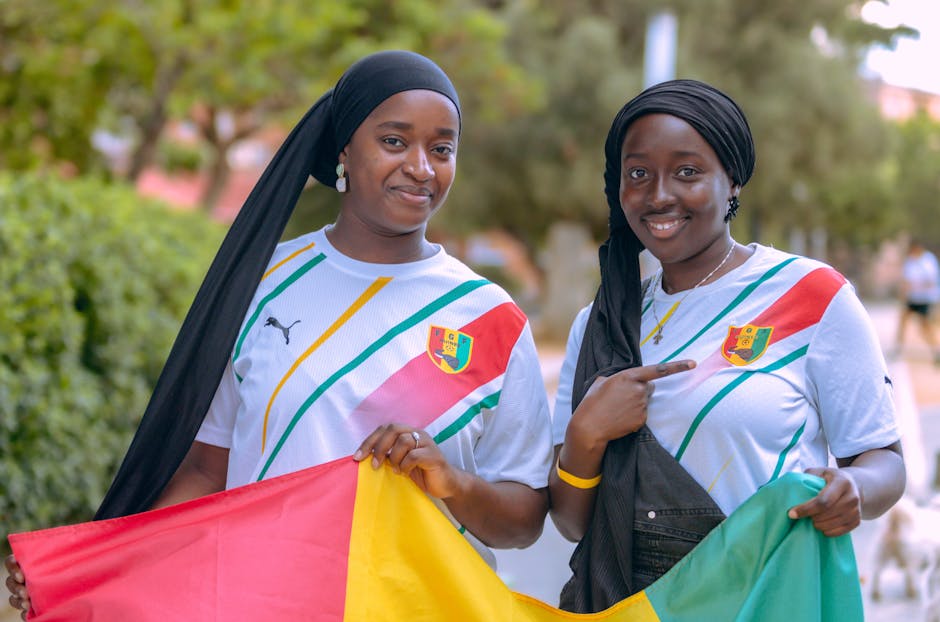 Two women wearing team jerseys hold a national flag outdoors, smiling and joyful.