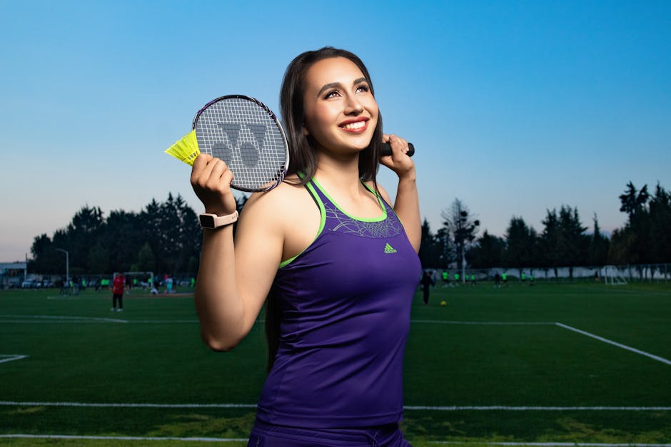 Smiling woman in athletic attire holding badminton racket on a sunny day outdoors.