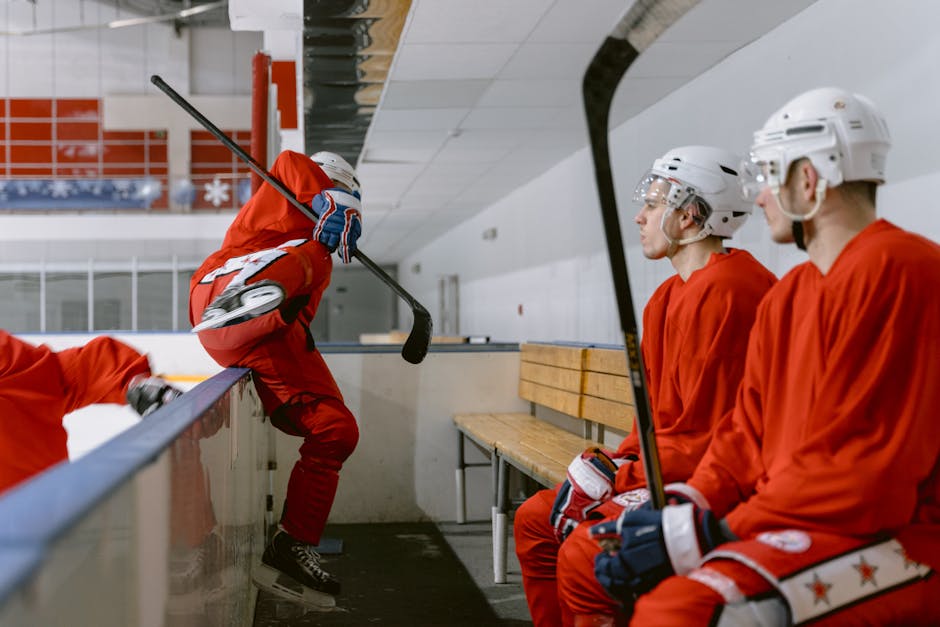 Ice hockey players in red uniforms sit and prepare on a rink bench.