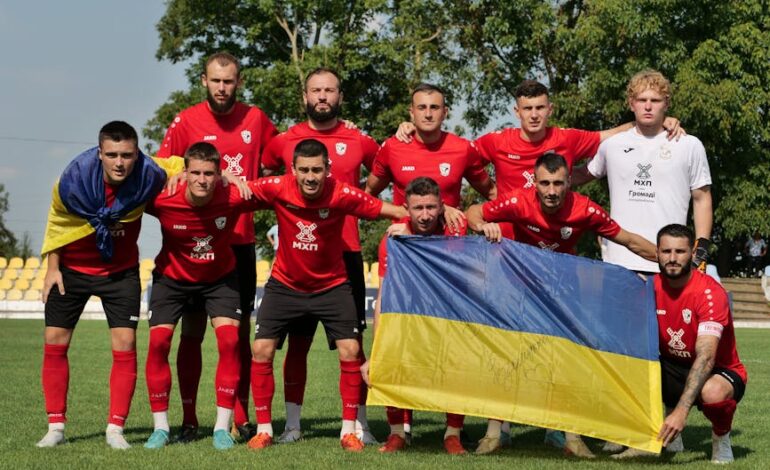 Soccer team in red jerseys posing with Ukrainian flag on a sunny day.