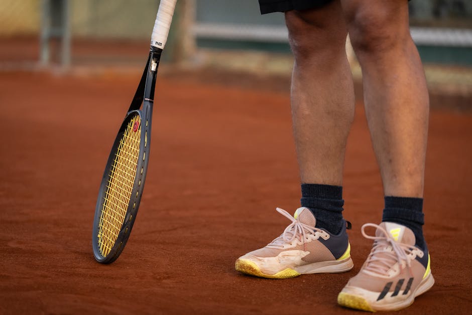 A close-up shot featuring a tennis racket and player's shoes on a clay tennis court.