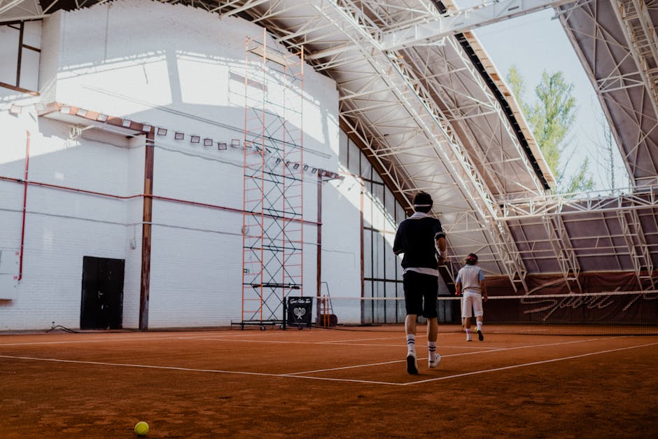 Two players engaging in a tennis match on an indoor clay court with natural lighting.