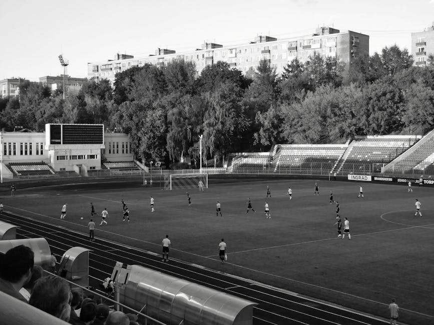 A black and white image of a soccer match taking place in an outdoor stadium with surrounding buildings.