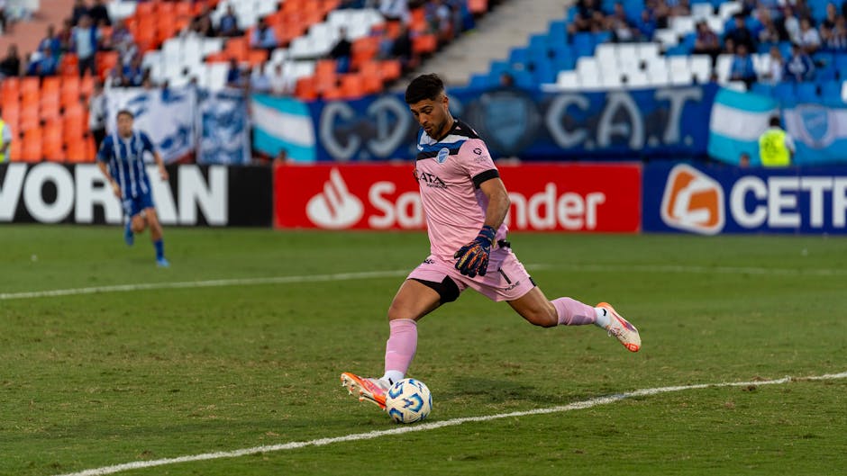 Goalkeeper in action kicking a football during an outdoor sports match, wearing pink kit