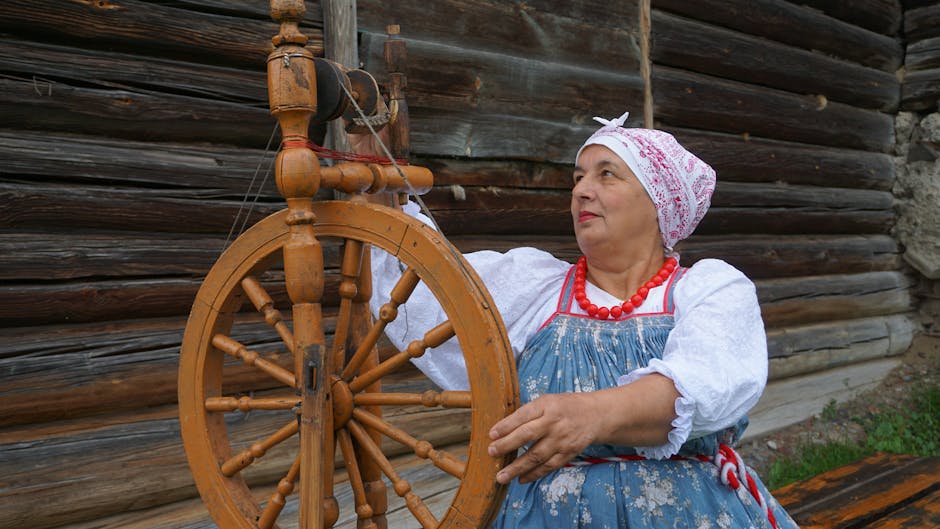 Elderly woman in traditional dress operating a wooden spinning wheel outdoors.