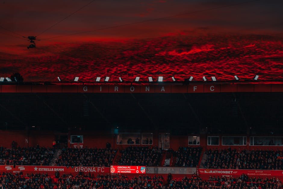 A stadium filled with fans under a dramatic red sunset sky during a Girona FC match.