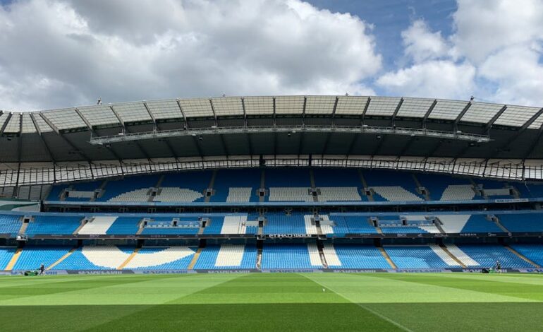 Etihad Stadium in Manchester with empty blue seats under a cloudy sky.