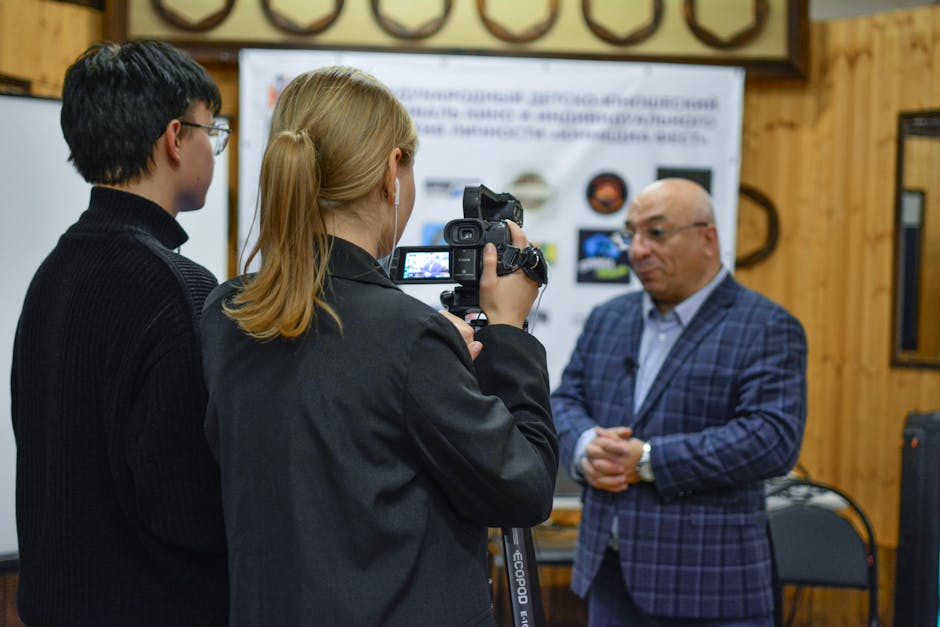 A journalist records an interview with a man using a camcorder indoors.