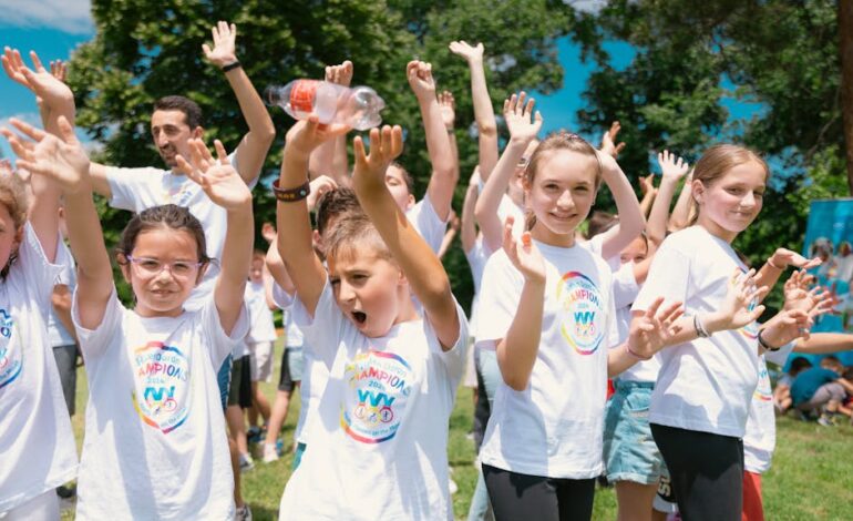 Group of cheerful children raising hands in a park during a sunny day, wearing t-shirts with event logos.