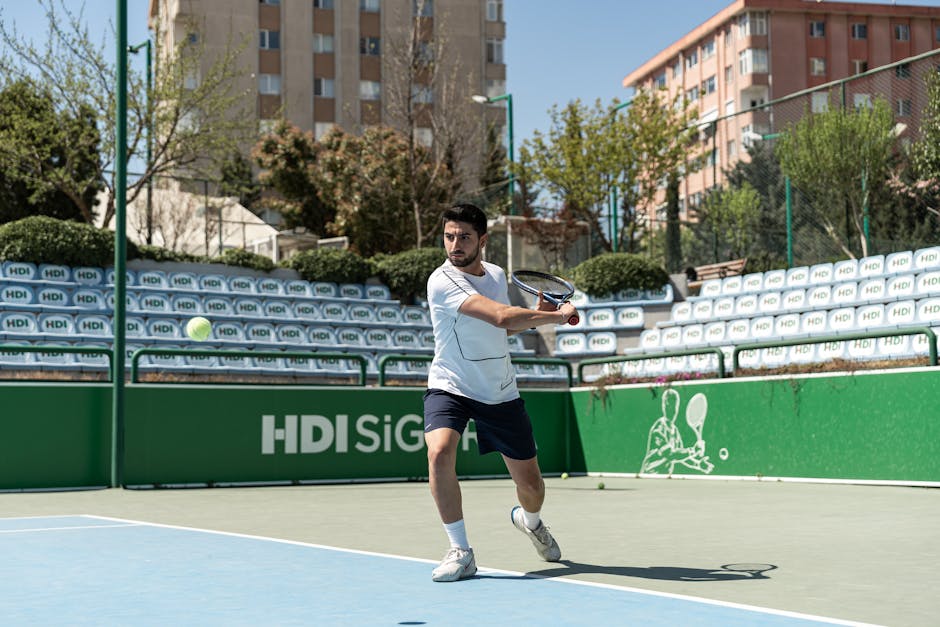 Male tennis player hitting a ball on an urban outdoor court during the day.