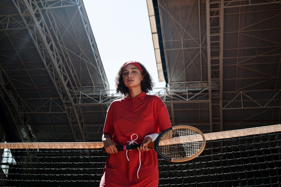 A woman in a red outfit stands with a tennis racket indoors, exuding vintage fashion vibes.