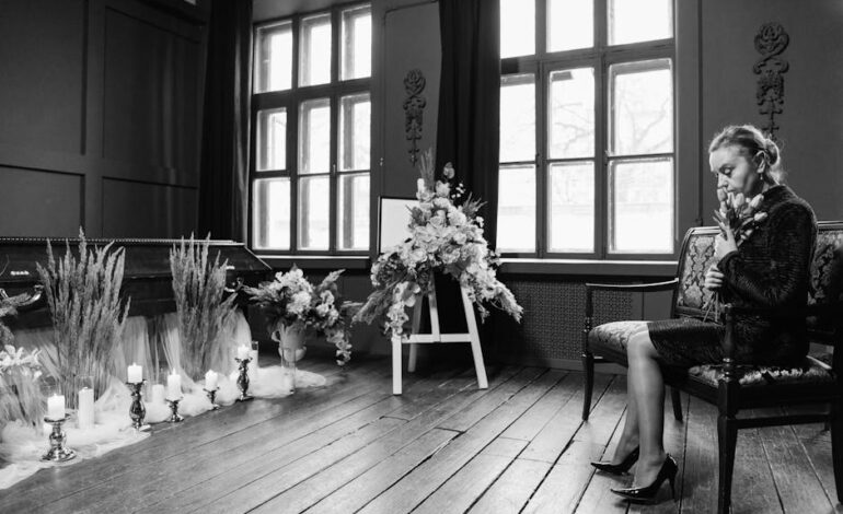 Grayscale funeral scene showing a woman in mourning by a coffin and floral arrangements.