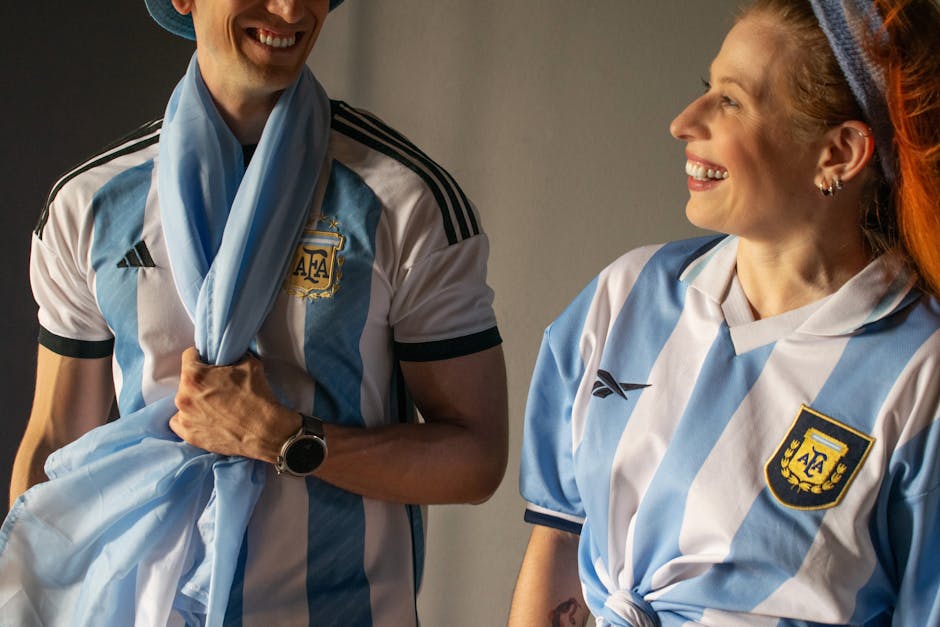 Two smiling fans wear official Argentina soccer jerseys and scarves indoors, showcasing team spirit.