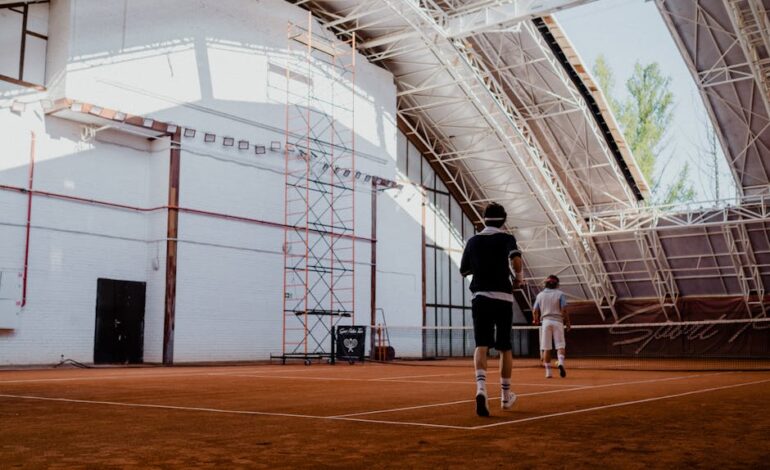 Two players engaging in a tennis match on an indoor clay court with natural lighting.