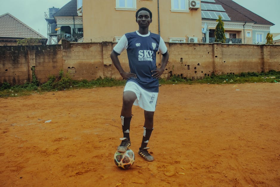 A teenage soccer player in uniform poses with a ball on a dirt field.